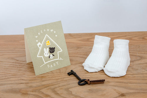 Welcome baby card with animal characters, white socks, and an old-fashioned key on a wooden table.